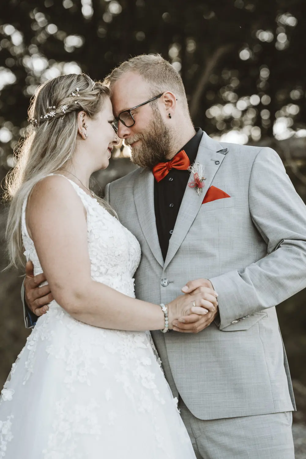 Eine Braut im weißen Spitzenkleid mit voluminösem Rock und floralem Kopfschmuck steht allein am steinigen Strand von Fehmarn. Umgeben von Treibholz, Wellen und Küstenwald blickt sie sanft nach unten – ein stiller, zeitloser Moment in Schwarz-Weiß-Fotografie voller Ruhe und natürlicher Hochzeitsästhetik aus Schleswig-Holstein