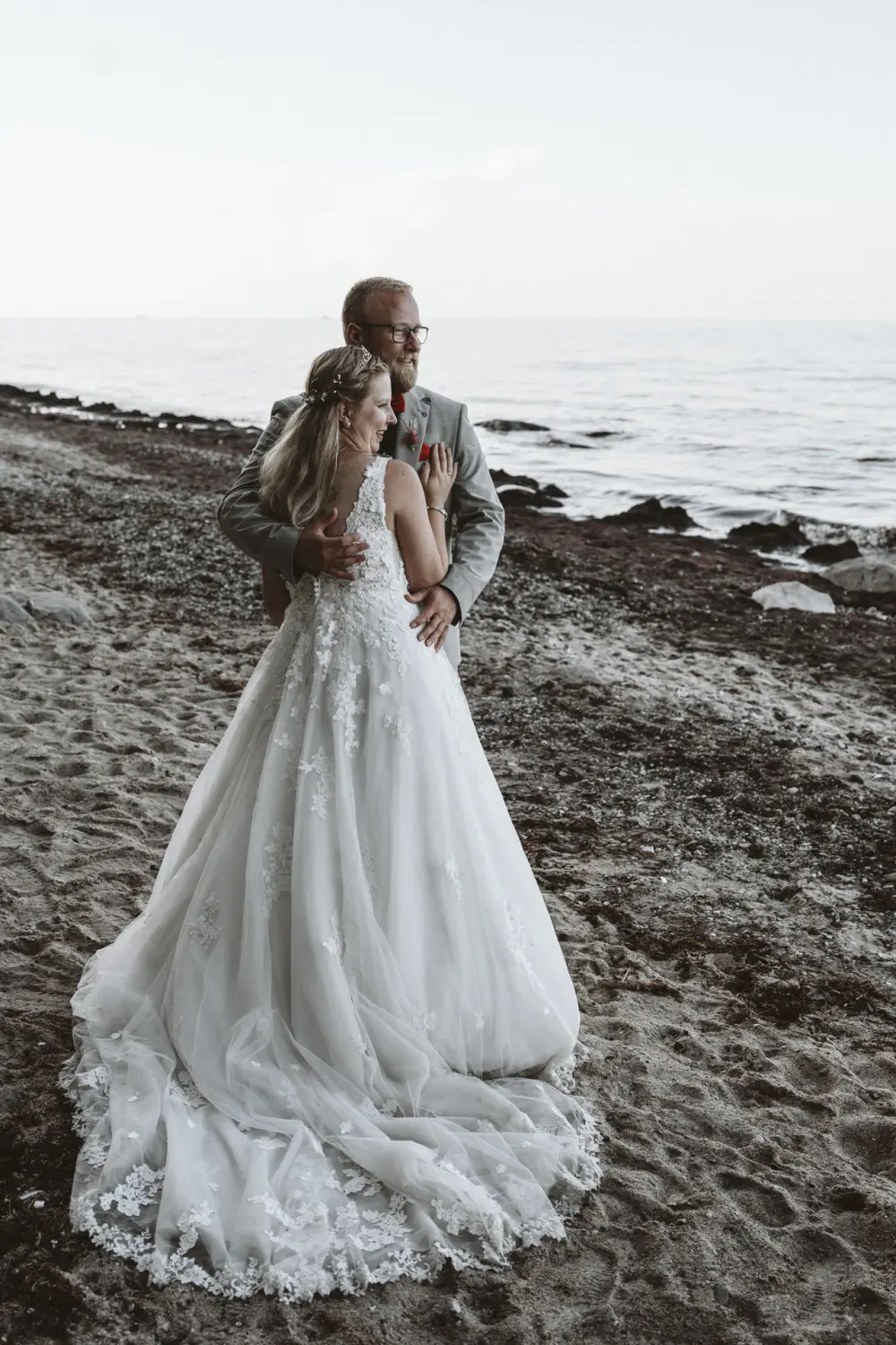 Ein Hochzeitspaar steht umarmt am Strand von Fehmarn: Die Braut in weißem Spitzenkleid mit langem Schleier und der Bräutigam im hellgrauen Anzug mit roter Blume blicken gemeinsam aufs Wasser. Ihr Stand auf Sand und Felsen, umgeben von Wellen und Himmel, zeigt einen ruhigen After-Wedding-Moment – natürliche Hochzeitsfotografie aus Schleswig-Holstein