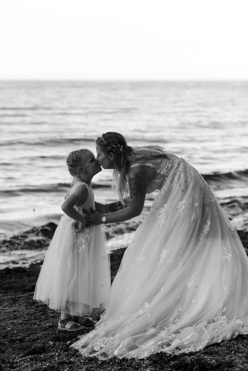 Eine Braut im weißen Spitzenkleid und ihre kleine Tochter  in ähnlicher Kleidung stehen gemeinsam am Ostseestrand von Fehmarn. Die Braut beugt sich liebevoll zu ihrem 6 Jahre alten Kind herab – ein inniger Moment voller Verbindung, eingefangen in natürlicher Hochzeitsfotografie aus Schleswig-Holstein