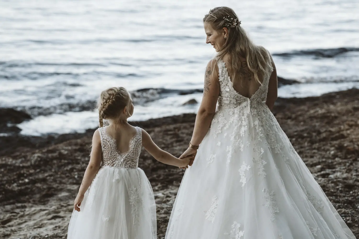 Eine Braut im weißen Spitzenkleid mit floraler Tiara und ein kleines Mädchen in einem ähnlichen Kleid stehen Hand in Hand am Ufer von Fehmarn. Beide blicken aufs Meer, umgeben von Wellen und Weite. Ein stiller After-Wedding-Moment voller liebevoller Verbindung und natürlicher Hochzeitsfotografie aus Schleswig-Holstein
