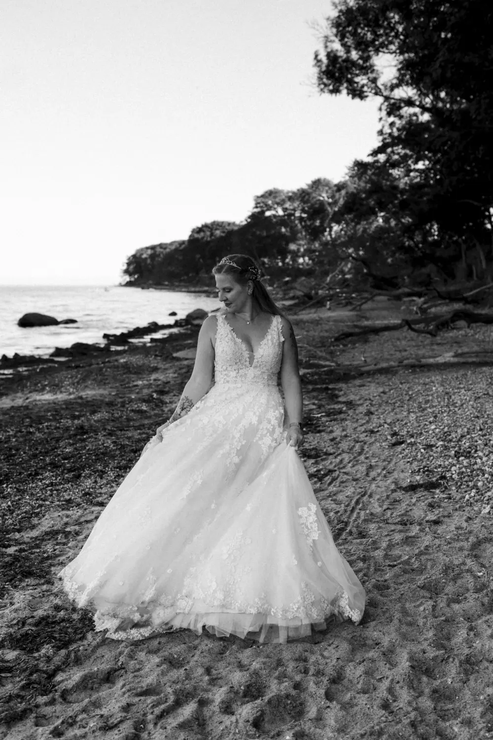 Eine Braut im weißen Spitzenkleid mit voluminösem Rock und floralem Kopfschmuck steht allein am steinigen Strand von Fehmarn. Umgeben von Treibholz, Wellen und Küstenwald blickt sie sanft nach unten – ein stiller, zeitloser Moment in Schwarz-Weiß-Fotografie voller Ruhe und natürlicher Hochzeitsästhetik aus Schleswig-Holstein