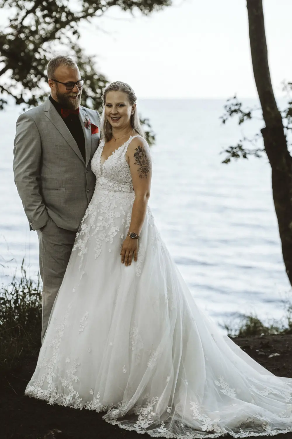 Braut und Bräutigam stehen eng beieinander am Katharinhof auf Fehmarn, direkt an der Ostsee. Der Bräutigam trägt einen grauen Anzug mit schwarzem Hemd und rotem Einstecktuch. Die Braut hat ein weißes Spitzenkleid und ein florales Tattoo auf dem Oberarm. Umgeben von Wasser, Wiesen und Bäumen wirkt das After-Wedding-Shooting wie ein stilles Küstenmärchen.