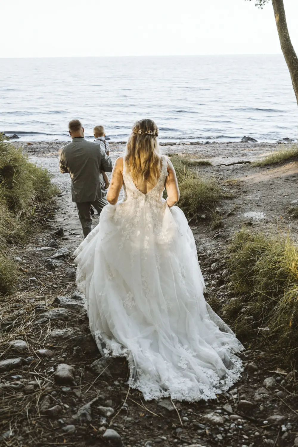 Ein Hochzeitspaar geht mit einem kleinen Kind in den Armen einen steinigen Pfad hinunter zum Strand von Fehmarn. Die Braut trägt ein weißes Spitzenkleid, der Bräutigam einen grauen Anzug mit roter Fliege. Ein inniger, natürlicher After-Wedding-Moment am Meer – emotionale Hochzeitsfotografie aus Schleswig-Holstein.