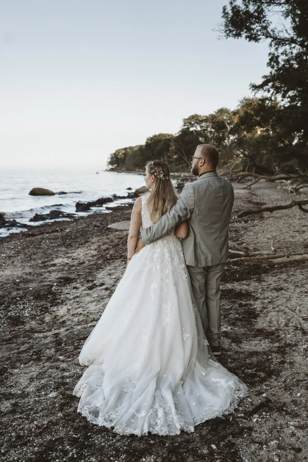 Ein Hochzeitspaar steht eng umschlungen am Sandstrand von Fehmarn: Die Braut im weißen Spitzenkleid mit floralem Haarschmuck, der Bräutigam im hellgrauen Anzug mit Brille und roten Akzenten – beide blicken gemeinsam aufs ruhige Wasser. Umgeben von Treibholz, Steinen und Küstenwald entsteht ein natürlicher After-Wedding-Moment voller Nähe und Hochzeitsfotografie aus Schleswig-Holstein.