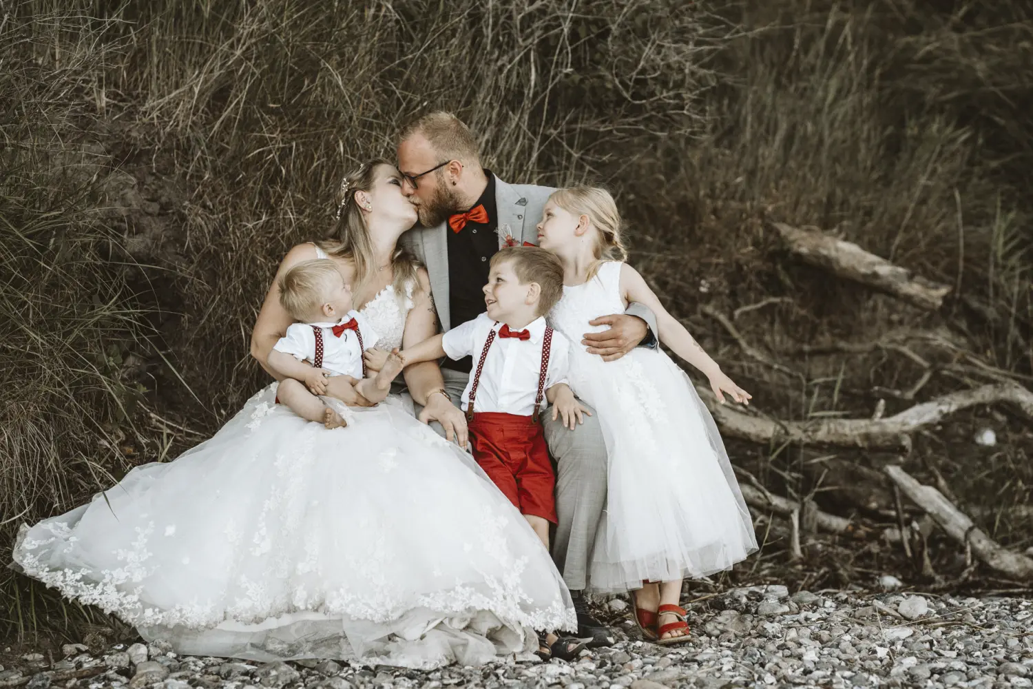 Eine Familie sitzt gemeinsam auf dem steinigen Boden am Ostseeufer von Fehmarn: Die Braut in weißem Spitzenkleid und der Bräutigam im grauen Anzug mit roter Fliege sind umgeben von ihren Kindern – ein Junge in weißem Hemd und roten Shorts mit Hosenträgern und ein Mädchen im weißen Kleid mit roten Sandalen. Ein liebevoller After-Wedding-Moment voller Nähe und natürlicher Hochzeitsfotografie in Schleswig-Holstein