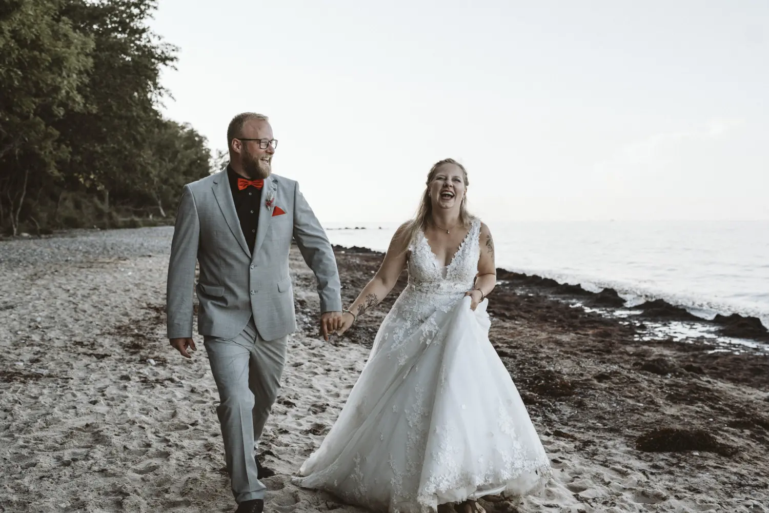 Ein Hochzeitspaar spaziert Hand in Hand am Strand von Fehmarn: Die Braut trägt ein weißes Spitzenkleid, der Bräutigam einen grauen Anzug mit roter Fliege. Gemeinsam in Bewegung, begleitet von Wellen und Küstenwald – ein natürlicher After-Wedding-Moment voller Leichtigkeit und Hochzeitsfotografie aus Schleswig-Holstein