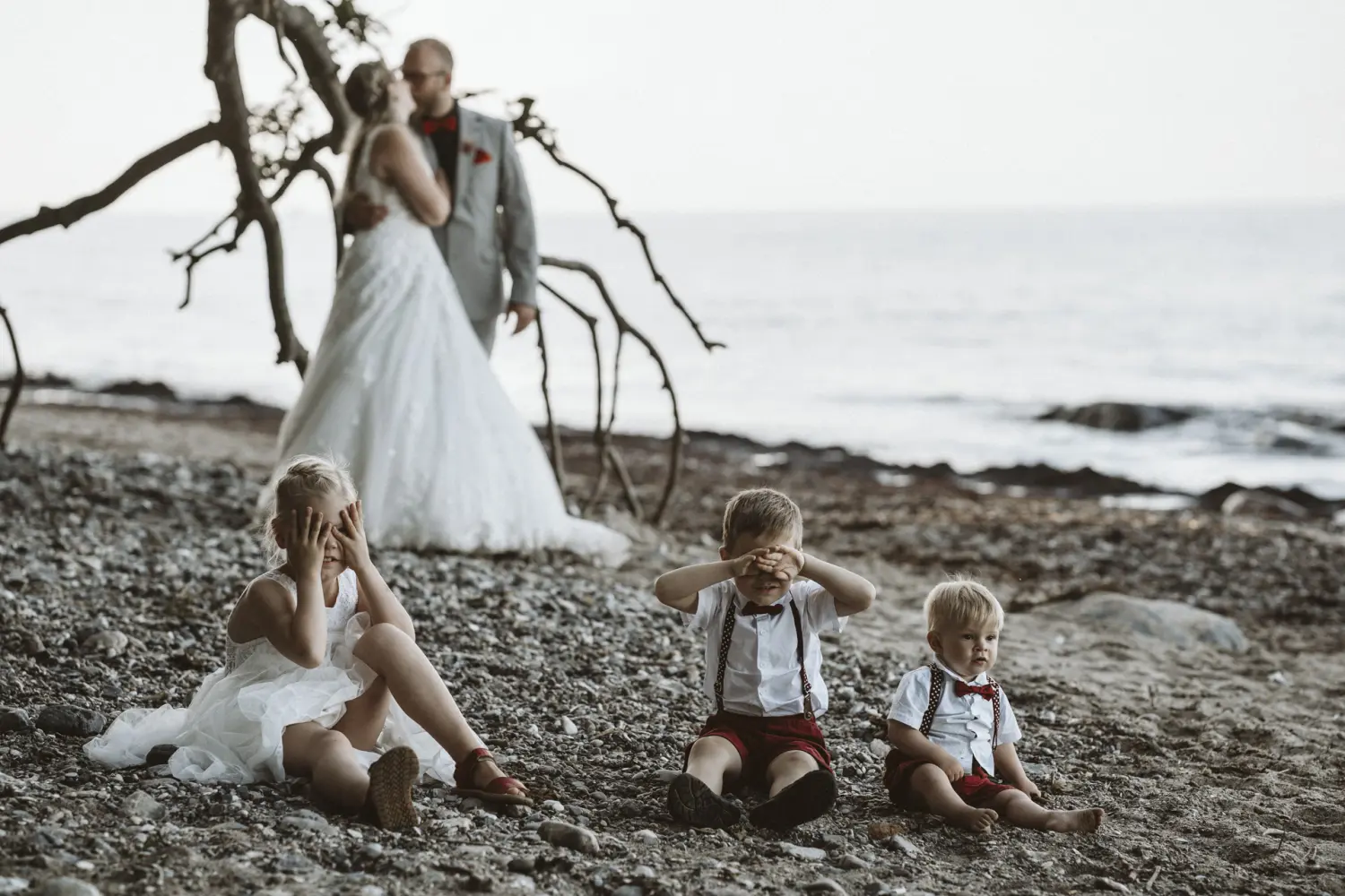 Ein Hochzeitspaar am Strand von Fehmarn steht eng umschlungen unter einem Baum: Die Braut trägt ein weißes Spitzenkleid, der Bräutigam einen grauen Anzug mit roter Fliege. Im Vordergrund sitzen drei Kinder auf den Steinen – zwei Jungen in weißen Hemden mit roten Shorts und eine kleine Brautjungfer im weißen Kleid. Ein natürlicher After-Wedding-Moment voller Familienliebe und Strandatmosphäre – Hochzeitsfotografie aus Schleswig-Holstein mit Herz.