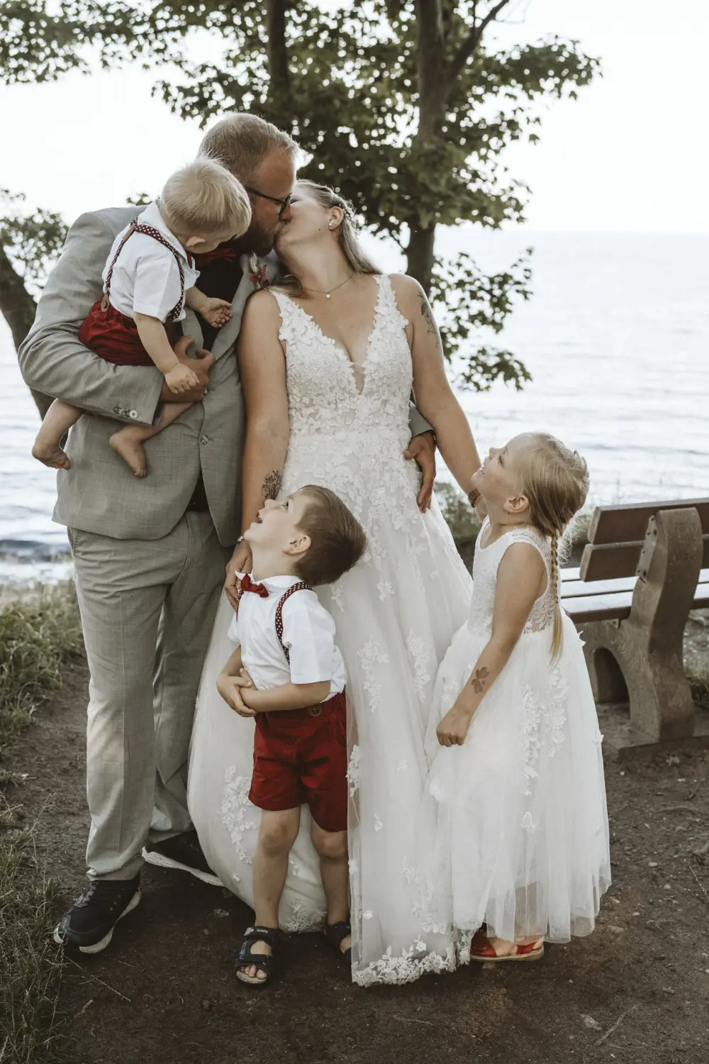 Ein Hochzeitspaar mit drei Kindern steht am Ufer von Fehmarn: Die Braut im weißen Spitzenkleid und der Bräutigam im grauen Anzug mit roter Fliege erleben gemeinsam mit ihren Kindern einen liebevollen After-Wedding-Moment – natürliche Hochzeitsfotografie in Schleswig-Holstein voller echter Emotionen.Hochzeitsfotografie in Schleswig-Holstein voller echter Emotionen.