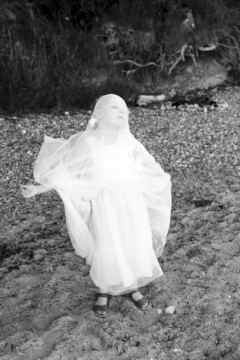 Ein kleines Mädchen steht allein am Sandstrand von Fehmarn. Sie trägt ein weißes Kleid, das sie sich spielerisch über den Kopf gezogen hat. Umgeben von Gras, Steinen und Treibholz wirkt sie wie in ihre eigene Fantasiewelt versunken – ein verträumter After-Wedding-Moment voller kindlicher Leichtigkeit in Schwarz-Weiß-Fotografie aus Schleswig-Holstein.