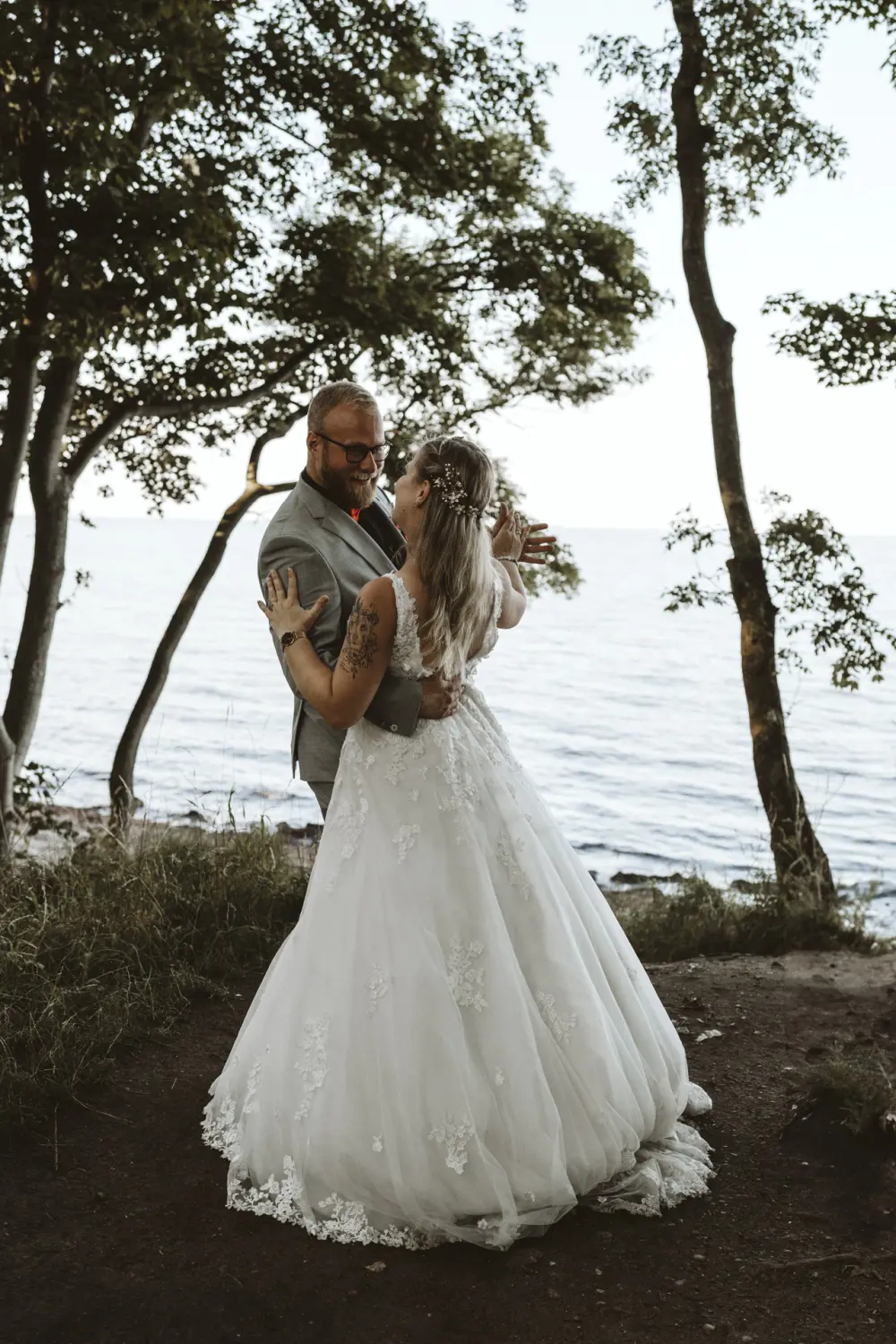 Die Braut im weißen Spitzenkleid und der Bräutigam im grauen Anzug mit roter Fliege tanzen am Ufer von Fehmarn – ein liebevoller After-Wedding-Moment voller Leichtigkeit und natürlicher Hochzeitsfotografie in Schleswig-Holstein.