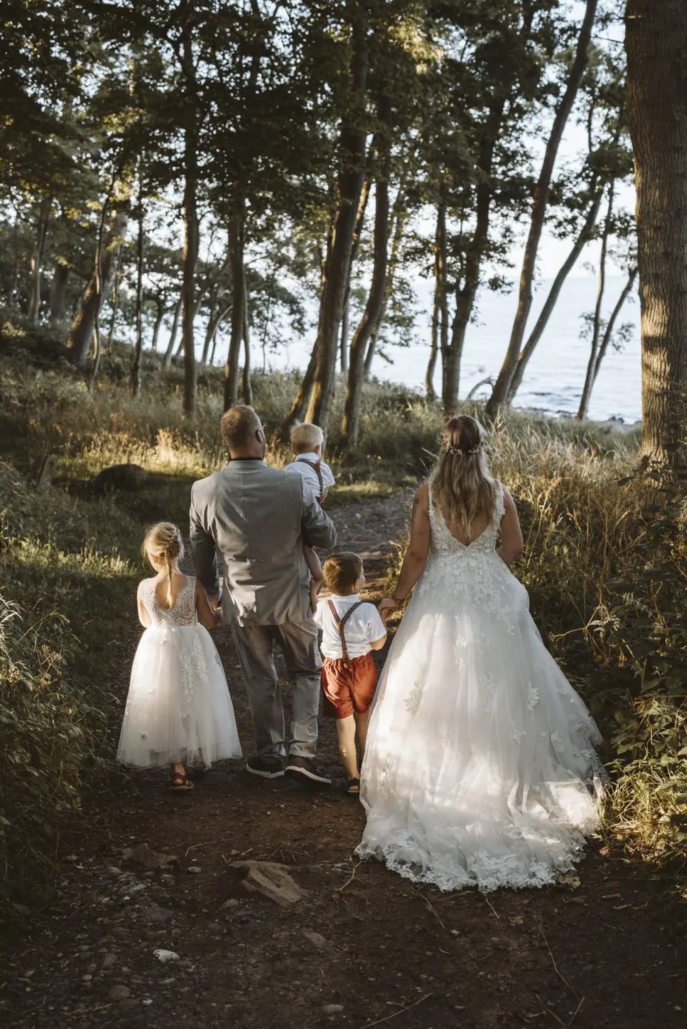 Eine Familie beim After-Wedding-Shooting spaziert durch den Wald in Richtung Ostseeufer von Fehmarn: Der Bräutigam in grauem Anzug trägt liebevoll das jüngste Kind, die Braut im weißen Spitzenkleid begleitet ihn mit zwei weiteren Kindern in festlicher Kleidung. Ein stiller, herzlicher Moment natürlicher Hochzeitsfotografie in Schleswig-Holstein.