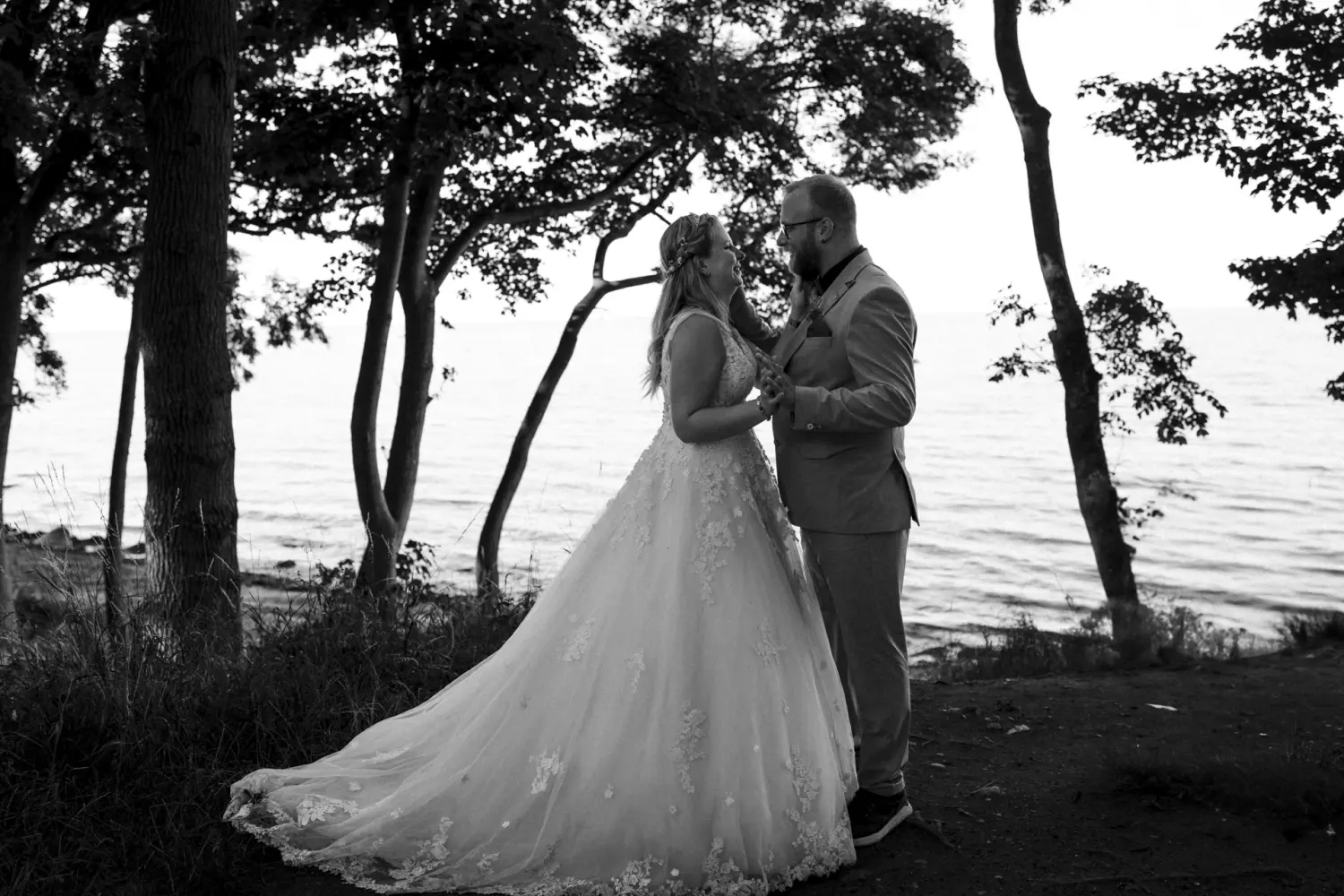 Die Braut im weißen Spitzenkleid und der Bräutigam im eleganten Anzug stehen eng umschlungen zwischen Bäumen am Ufer von Fehmarn – ein inniger, zeitloser Hochzeitsmoment in natürlicher Schwarz-Weiß-Fotografie aus Schleswig-Holstein